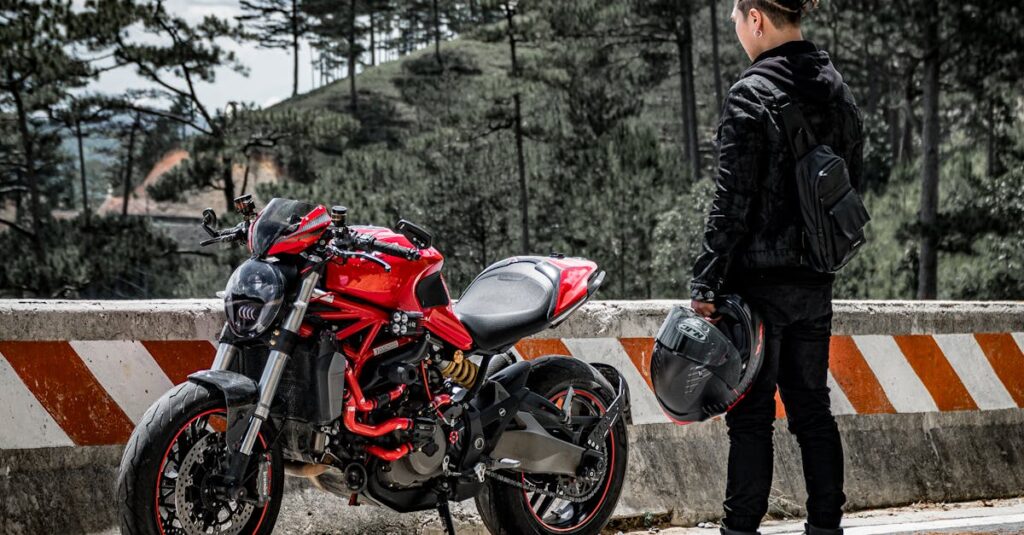 A man stands in front of a red sport motorcycle on a mountain road, surrounded by trees.